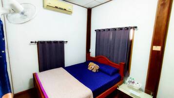 A small, single-bed room with a wooden headboard, a bedside table with a lamp, and an air conditioning unit mounted on the wall.