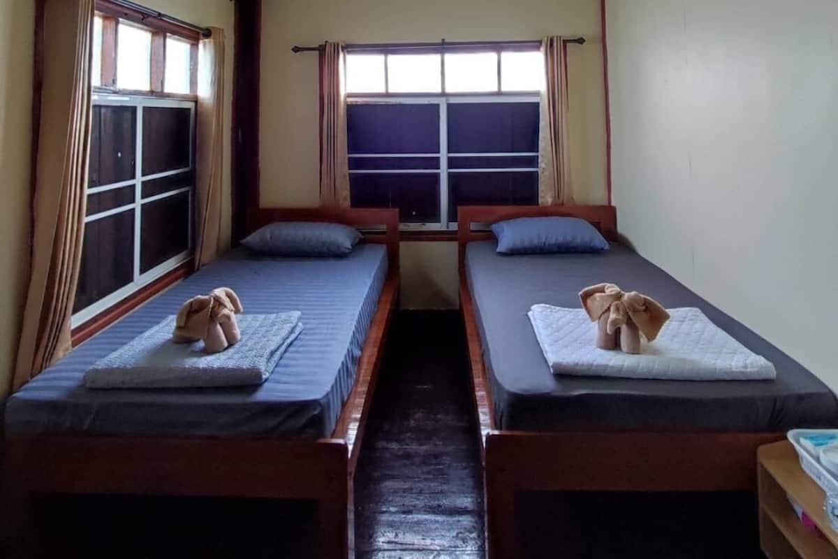 A room with two single beds, a window with curtains, an air conditioning unit, and a wooden floor.