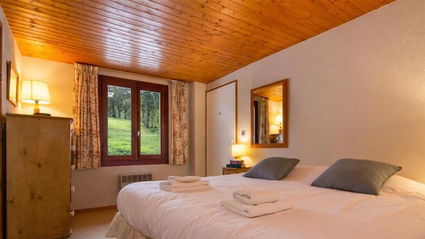 Een slaapkamer met een houten plafond, een bed met kussens en opgevouwen handdoeken, een dressoir, een raam met gordijnen en een spiegel aan de muur.