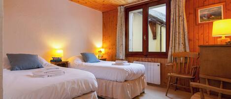 Una habitación con dos camas, paredes de madera, una ventana con cortinas, una lámpara y una silla.