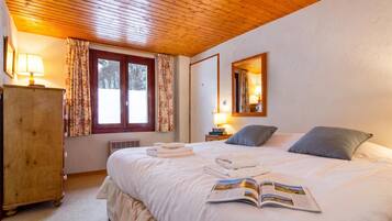 Une chambre d’hôtel avec un plafond en bois, un lit recouvert de draps blancs, une table de chevet avec une lampe, un miroir au mur et une fenêtre avec des rideaux.