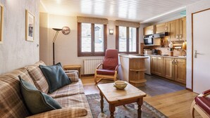 Een gezellige woonkamer met een bank, een rode fauteuil, een houten salontafel en een keukenhoek met houten kastjes en een magnetron.