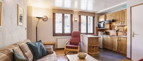 A cozy living room with a sofa, a red chair, a wooden coffee table, and a kitchen area with wooden cabinets and a microwave.