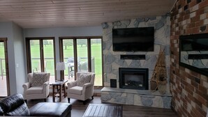 Un salon chaleureux avec une cheminée en pierre, des sièges confortables et de grandes fenêtres donnant sur un paysage verdoyant.