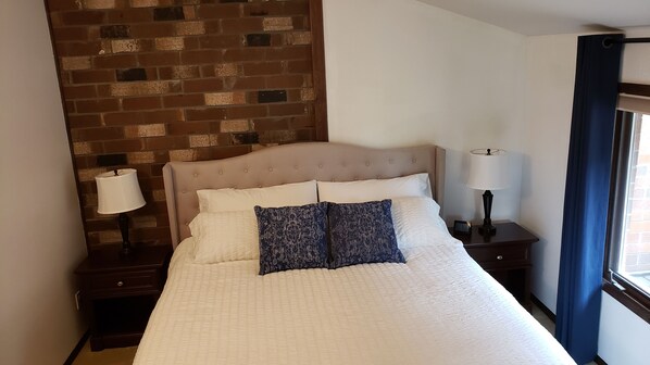 A hotel room with a brick wall, a bed with white and patterned pillows, two bedside tables with lamps, and a window with curtains.
