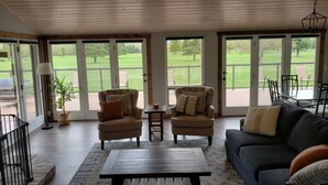 Un salon spacieux, doté d’un canapé foncé, de deux fauteuils, d’une table basse et offrant une vue sur une pelouse verdoyante grâce à de grandes portes-fenêtres coulissantes.