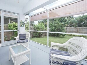 Un portico coperto con mobili in vimini e un tavolo in vetro, affaccia su un giardino recintato da una staccionata di legno e ricco di vegetazione.