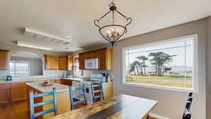 A kitchen with wooden cabinets, a central island with blue chairs, a dining table, and a large window offering a view of trees and a road.