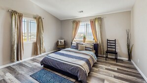 A bedroom with a large bed, two windows with curtains, a ceiling fan, and a view of the city.