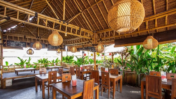 An outdoor restaurant with wooden tables and chairs, bamboo ceiling, and hanging lanterns.
