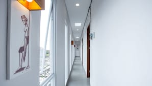 A long, narrow hallway with white walls and a grey floor, featuring a large window on the left and a door on the right.