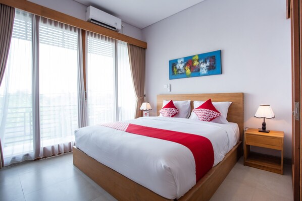 A hotel room with a large bed, a wooden headboard, a nightstand with a lamp, a painting on the wall, and a window with curtains. - OYO 1207 Pondok 789 (Munggu)