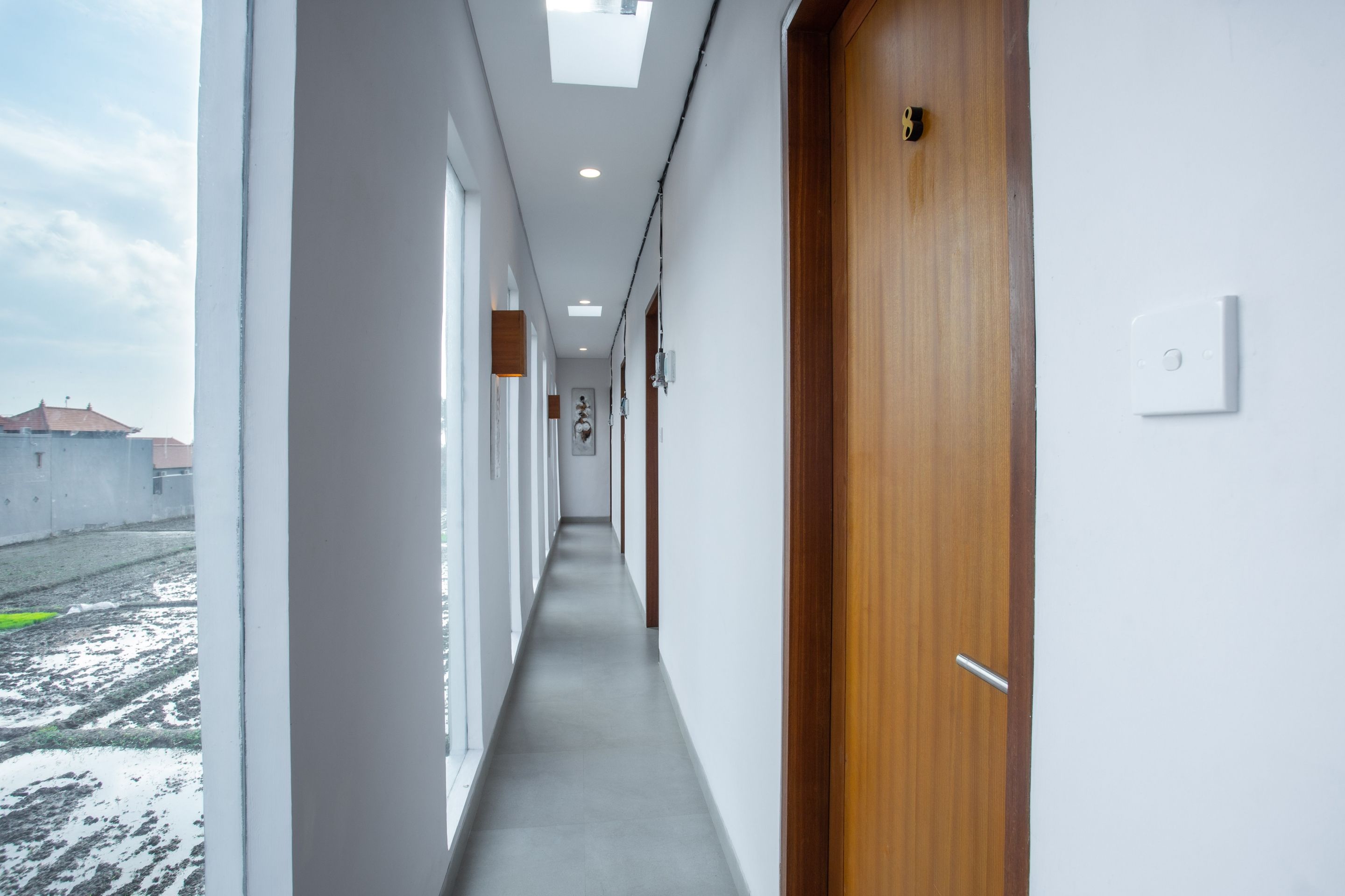 A corridor with a wooden door on the right, white walls, and a view of buildings outside.