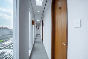 Een gang met een houten deur aan de rechterkant, witte muren en uitzicht op gebouwen.