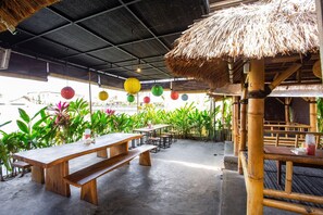 An outdoor dining area with wooden benches and tables, surrounded by greenery and thatched-roof structures.