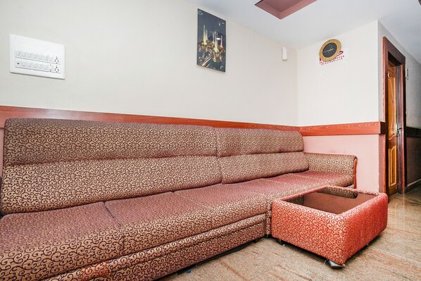 A patterned sofa with a matching ottoman in a room with a white wall, a clock, and a door. - OYO 436 Emirates Suites (Bengaluru)