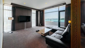 A modern living room with a large flat-screen TV, a sofa, a coffee table, and a view of the sea through large windows. - Hotel JCS Yeosu (Yeosu)