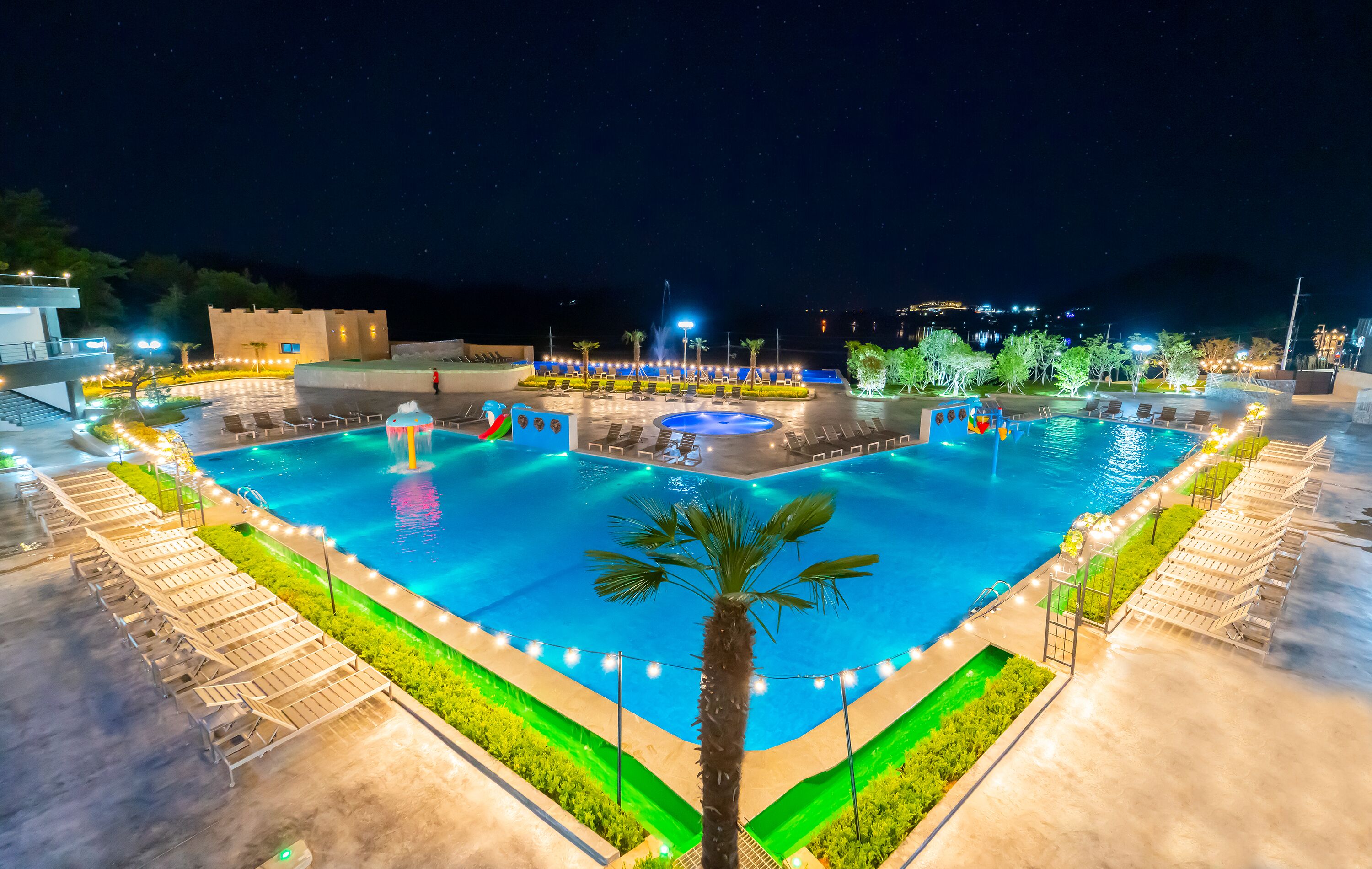 A large outdoor swimming pool with multiple lounge areas and a central water feature, surrounded by well-lit pathways and greenery.