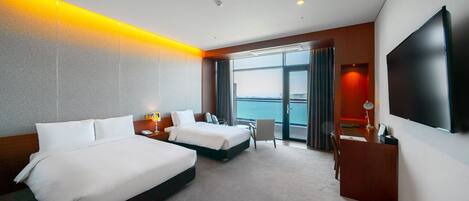 A hotel room with two beds, a desk, a chair, a TV, and a large window with a view of the sea.