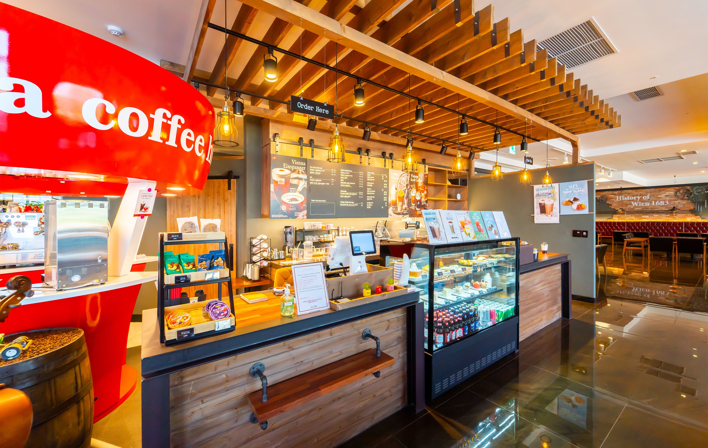 Una cafetería con un letrero rojo, un tablero de menú, una caja registradora y una vitrina con pasteles.
