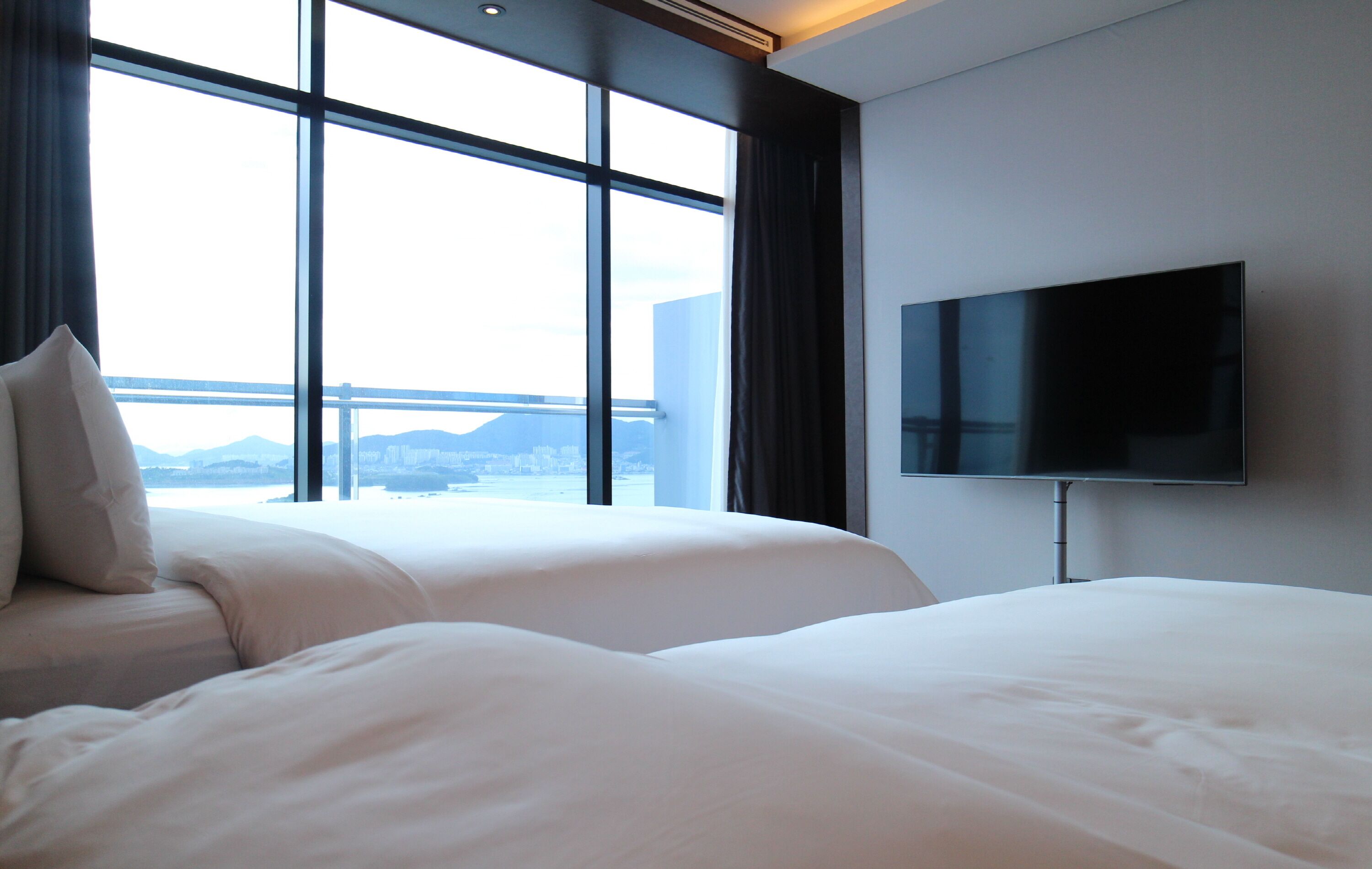 Quarto de hotel com duas camas, uma janela grande com vista para a cidade e uma televisão de tela plana fixada na parede.