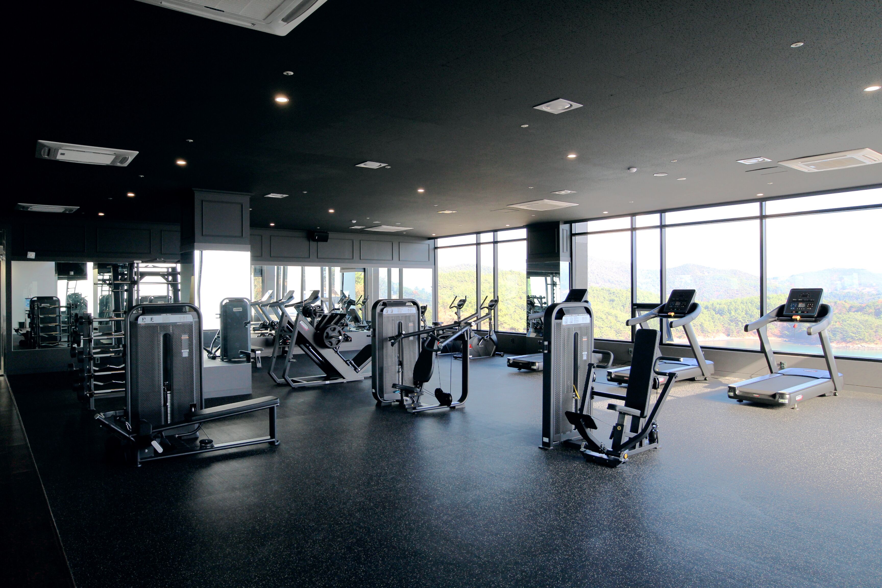 A modern gym with various exercise machines, large windows offering a view of the outdoors, and a spacious layout.