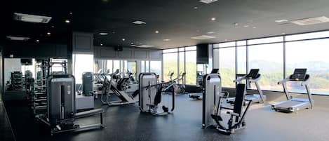 A modern gym with various exercise machines, large windows offering a view of the outdoors, and a spacious layout.