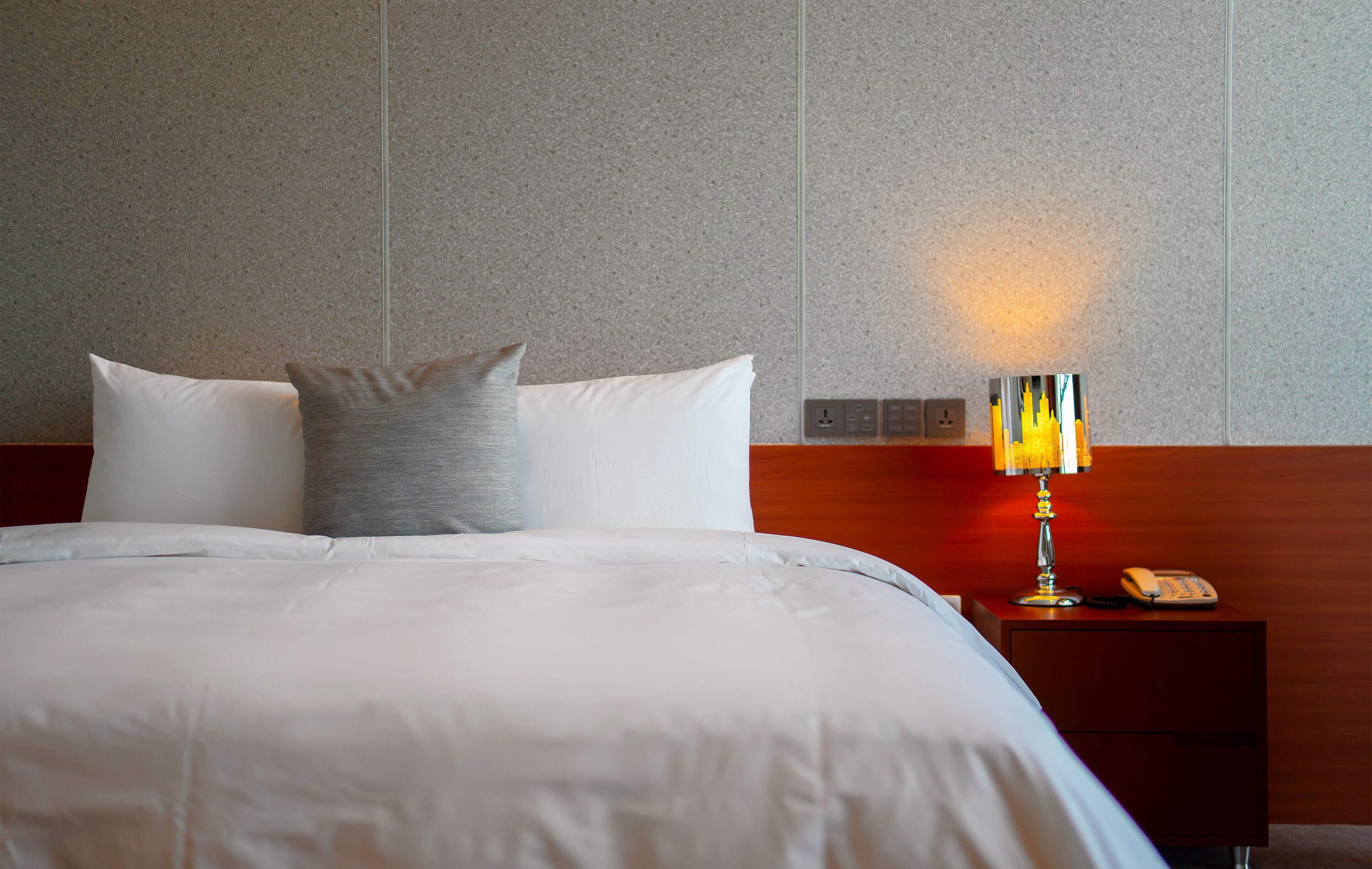 A neatly made bed with white bedding and a gray pillow, a bedside table with a lamp, and a phone.