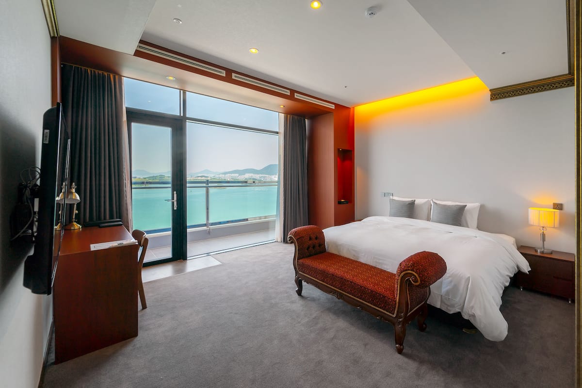 A hotel room with a large bed, a desk, a chair, a TV, and a view of the sea.