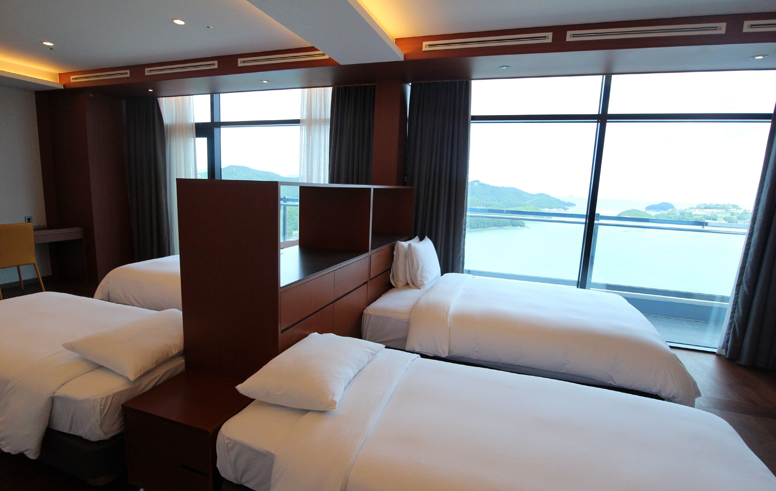 A hotel room with two beds, a desk, and a large window offering a view of a body of water and mountains.