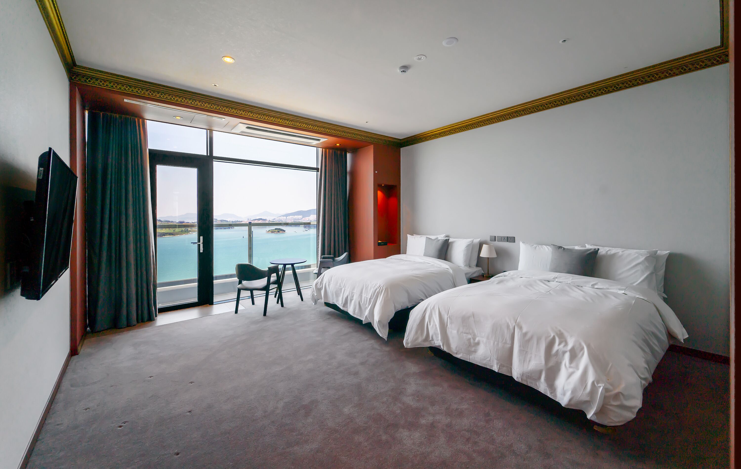 Quarto de hotel com duas camas, televisão, escrivaninha e vista para o mar.