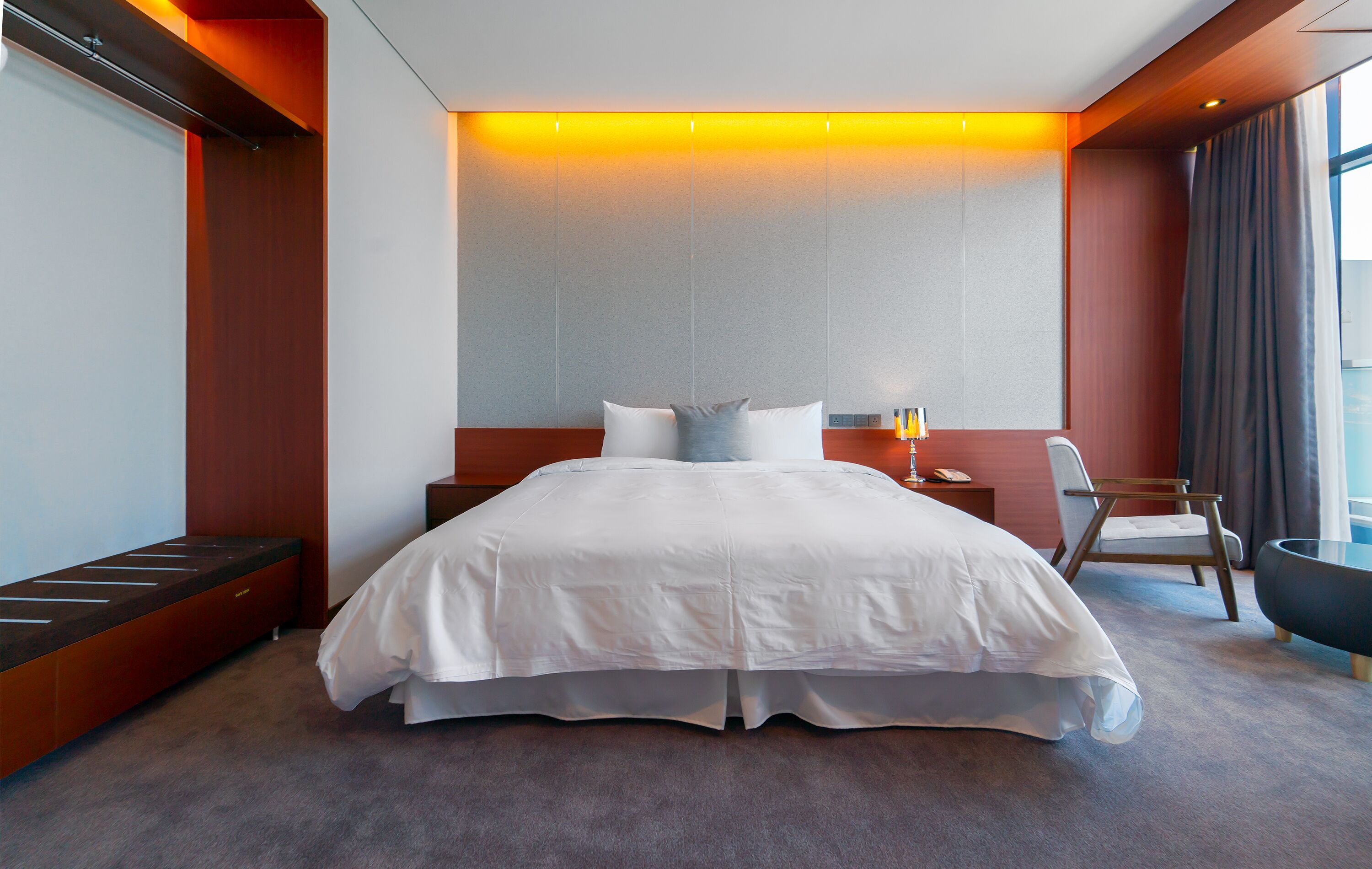 Quarto de hotel moderno com uma cama grande, mesa de cabeceira, uma cadeira e vista para a cidade.