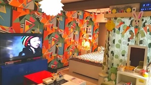 A modern living room with a TV, a sofa, a coffee table, and a bed with a colorful geometric wall design. - Chintai Navi Hostel (Osaka)