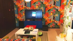 A living room with a TV, a coffee table with books, a sofa, and a colorful geometric wall pattern. - Chintai Navi Hostel (Osaka)