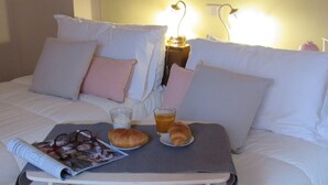 Een net opgemaakt bed met witte lakens, een nachtkastje met een tijdschrift, een croissant, een glas sinaasappelsap en een fles melk.