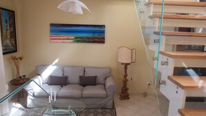 Een woonkamer met een bank, een glazen bijzettafeltje, een schilderij van een strand, een lamp en een trap.