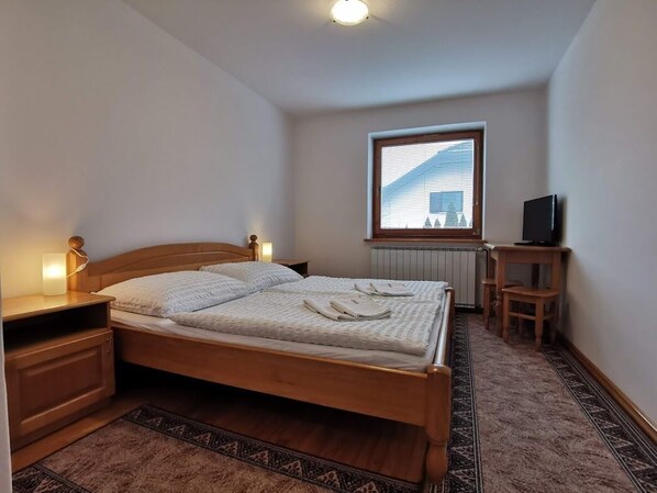 Un dormitorio con una cama de madera, lámparas de noche, una mesita de madera, un televisor sobre un mueble y una ventana con persianas.