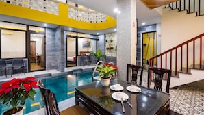 A modern indoor pool area with a dining table set for two, surrounded by chairs, and a staircase leading to an upper level. - The Lit Villa (Da Nang)