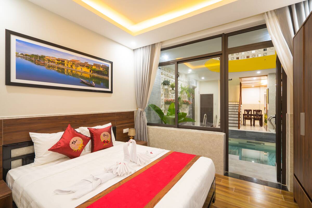 A hotel room with a bed, a painting, a window with curtains, and a view of a pool and dining area.