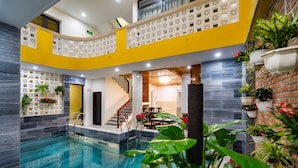 A modern indoor swimming pool with a curved yellow and white design, surrounded by potted plants and a brick wall. - The Lit Villa (Da Nang)