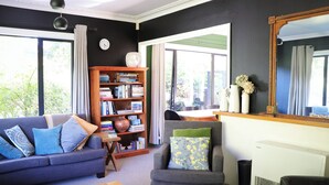 A living room with a sofa, armchair, and bookshelf, a dining area with a table and chairs, and a large window with curtains. - Light filled upstairs home minutes from central Hamilton (Claudelands)