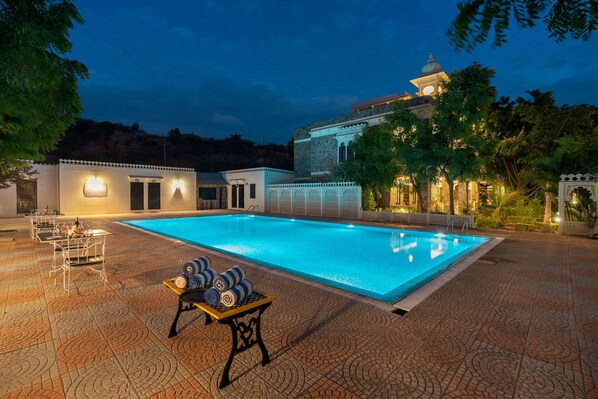 A swimming pool with a lounge area, a dining setup, and a building with traditional architecture. - The Udai Bagh (Mavli)