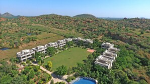 A resort with multiple buildings arranged around a central lawn, set in a hilly landscape with natural vegetation. - The Udai Bagh (Mavli)