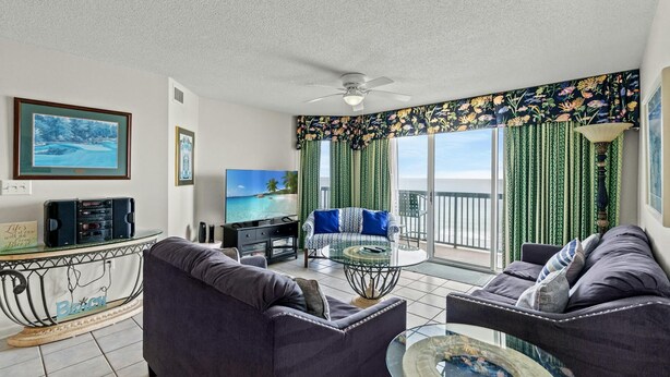 A living room with a sofa, a coffee table, and a TV. There is a balcony with a view of the ocean.