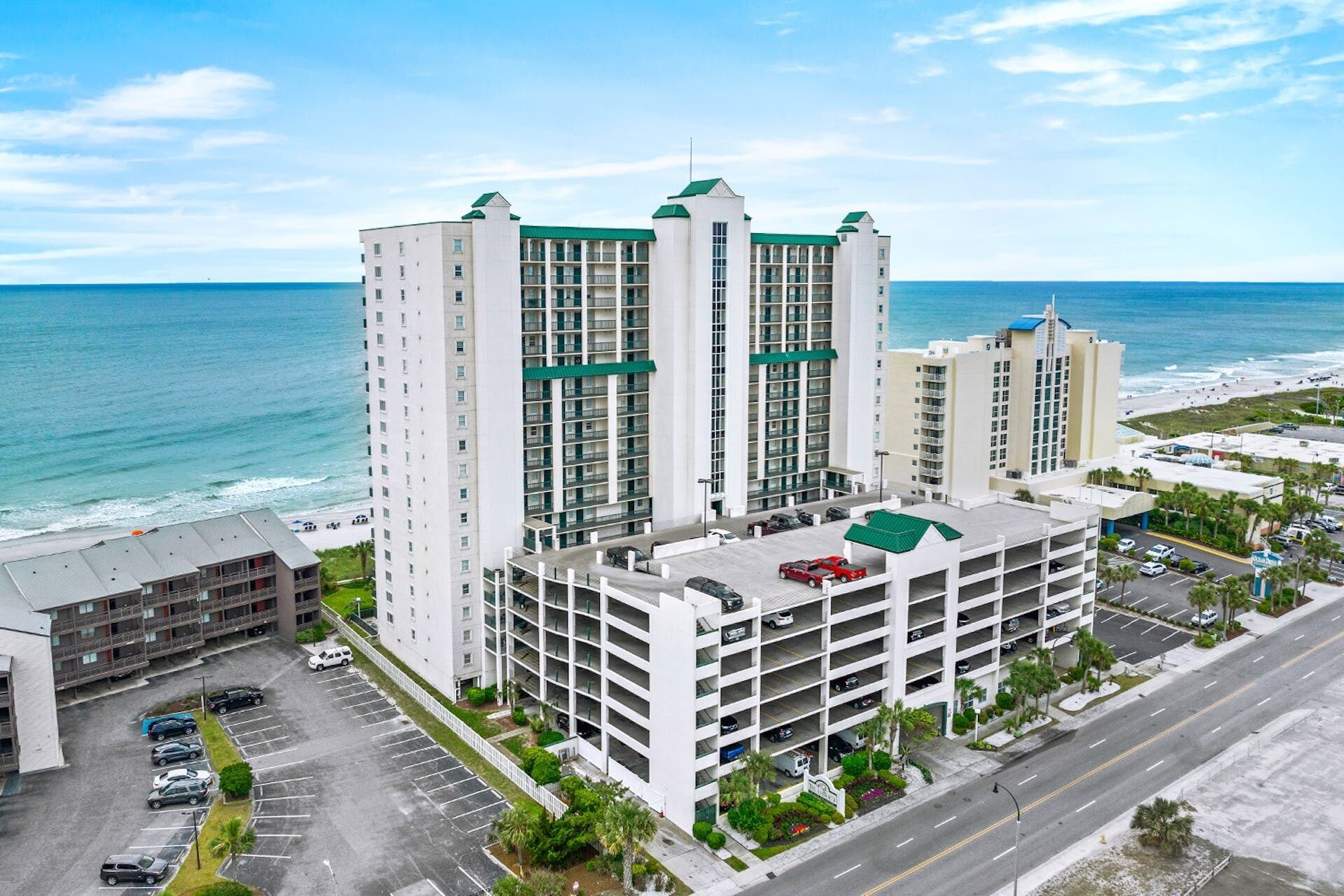 A multi-story hotel with a rooftop pool, located near a beach with a parking lot in front.