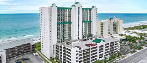 A multi-story hotel with a rooftop pool, located near a beach with a parking lot in front.
