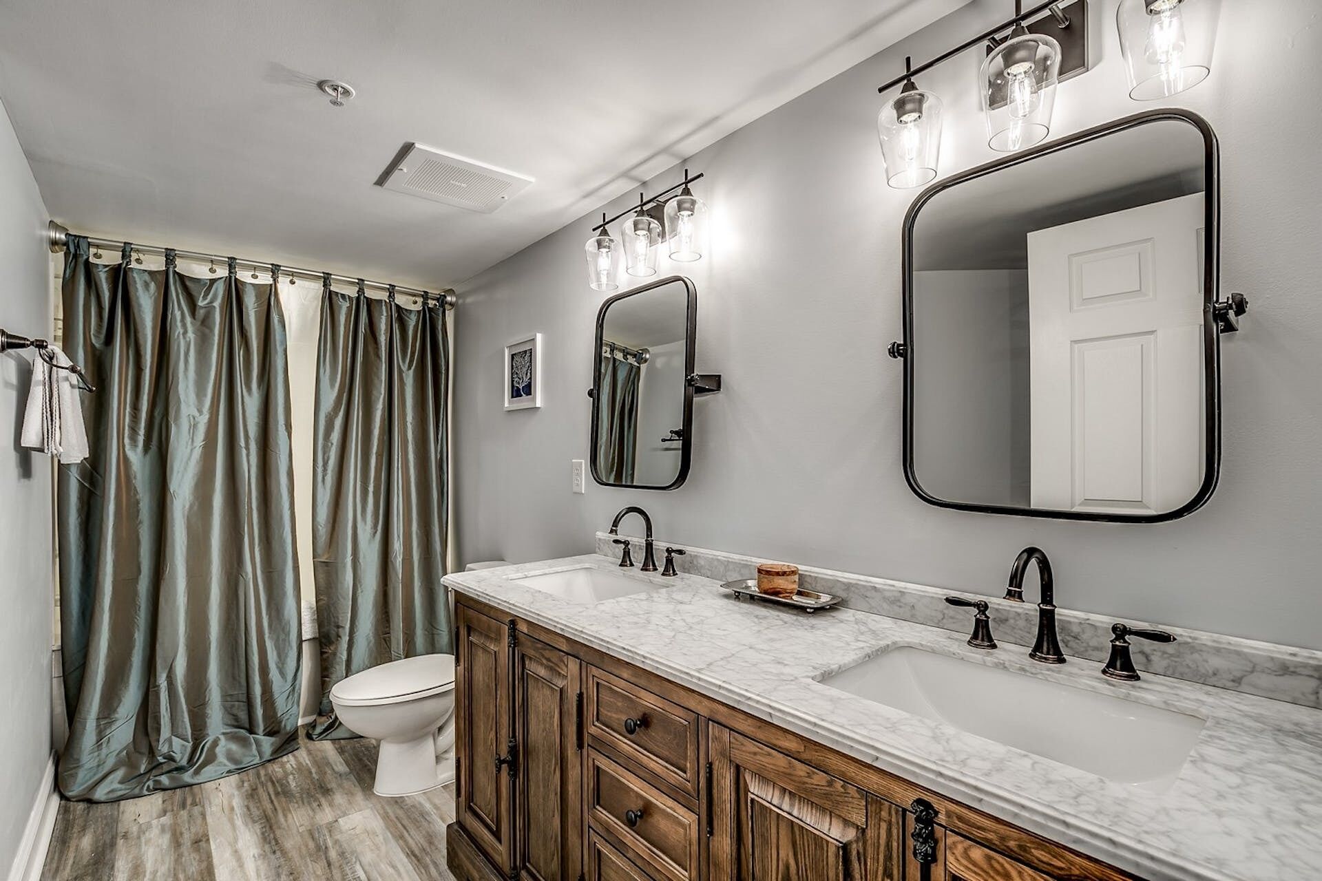A bathroom with a shower curtain, a toilet, a sink with a marble countertop, and two mirrors.