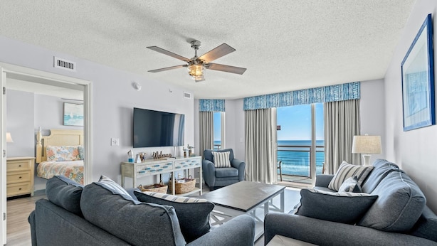 A modern living room with a large flat-screen TV, grey sofas, a coffee table, and a ceiling fan.
