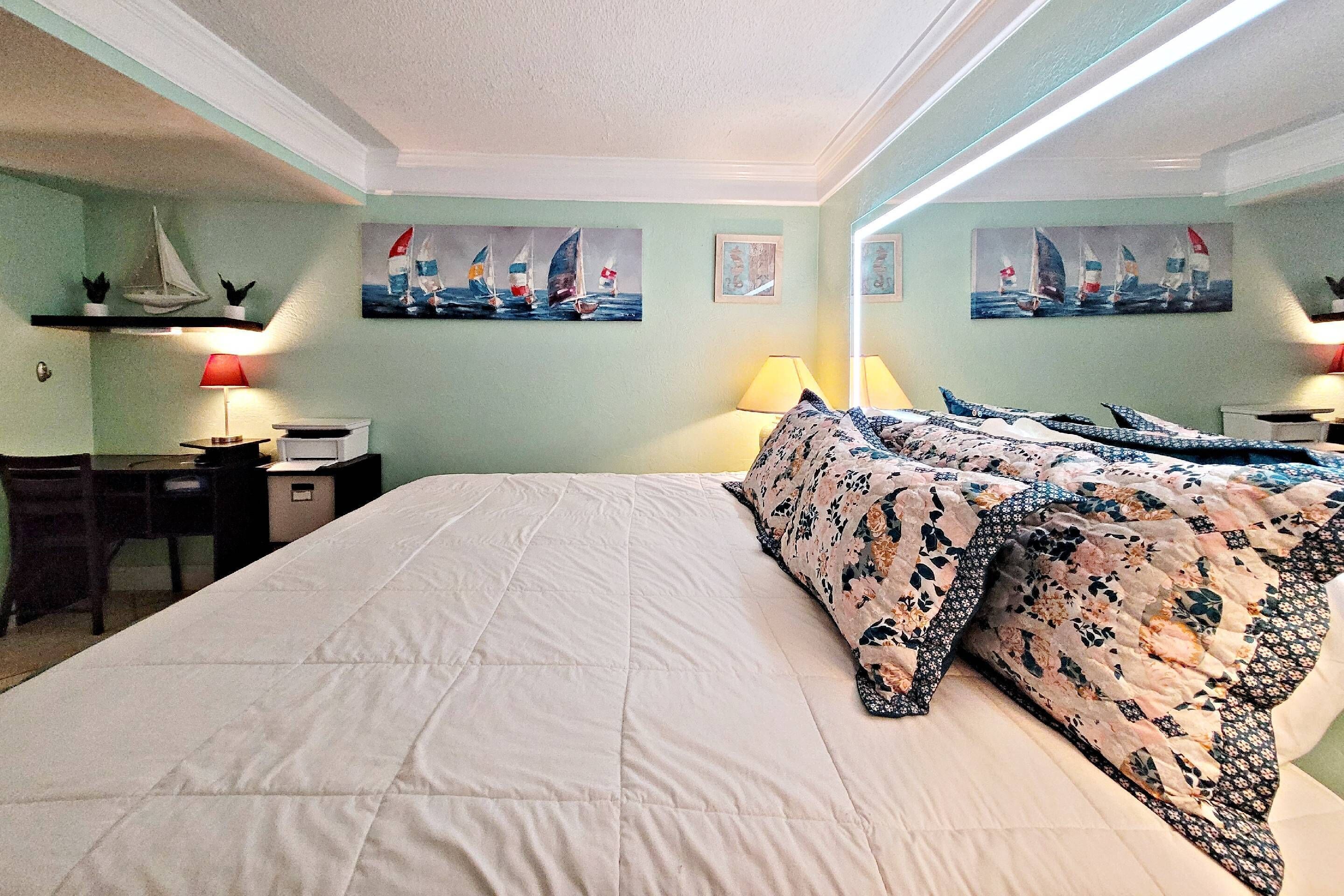 Un dormitorio con una cama grande, dos mesitas de noche con lámparas, un escritorio y una silla. Hay tres cuadros en la pared que representan veleros.