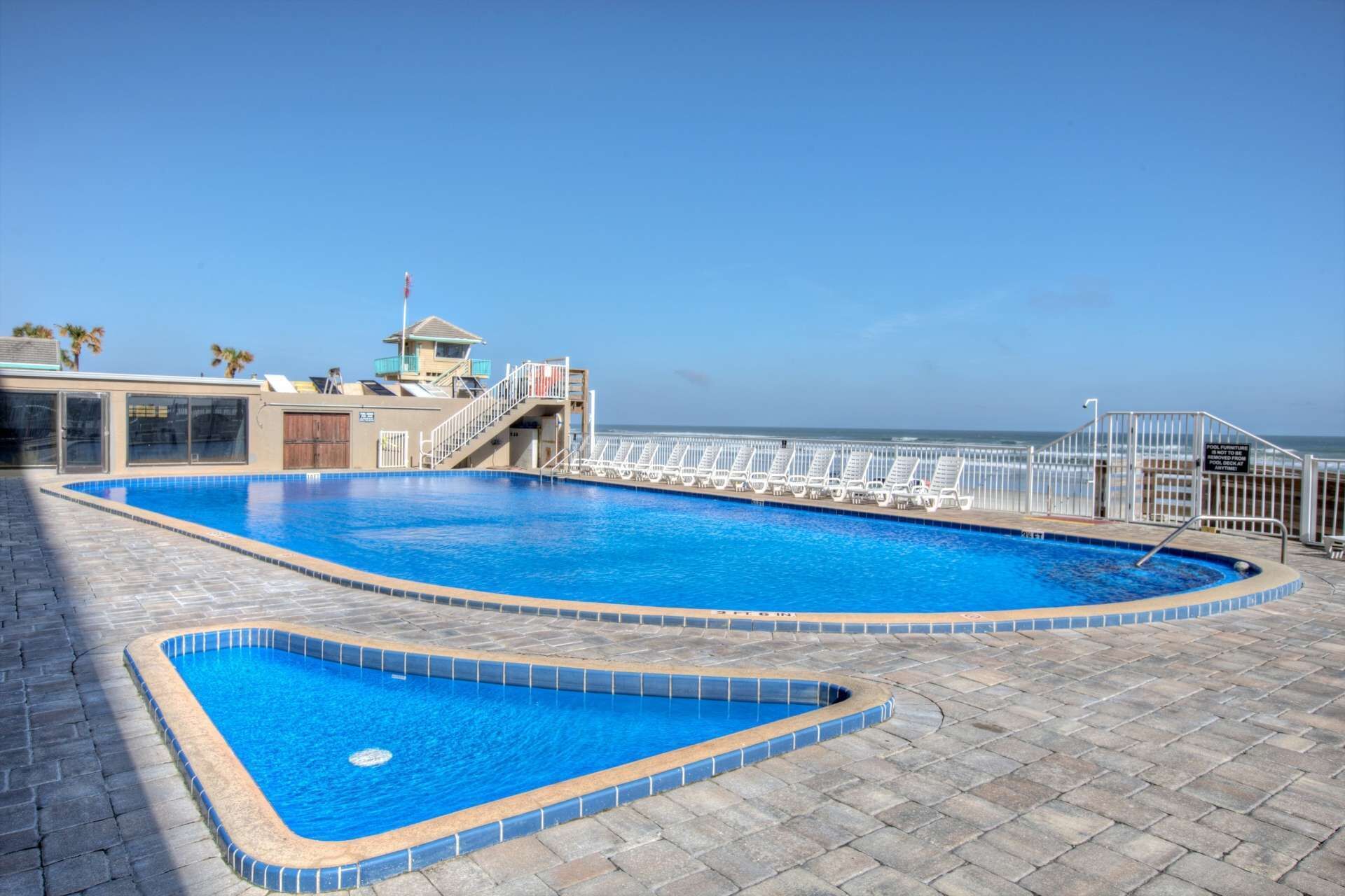 A rectangular swimming pool with a lifeguard tower, a deck with lounge chairs, and a view of the ocean.